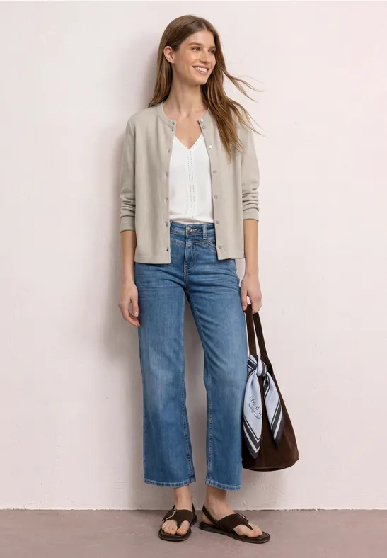 Vrouw met beige vest, witte blouse, wijd jeans, zwarte sandalen en een bruine tas.
