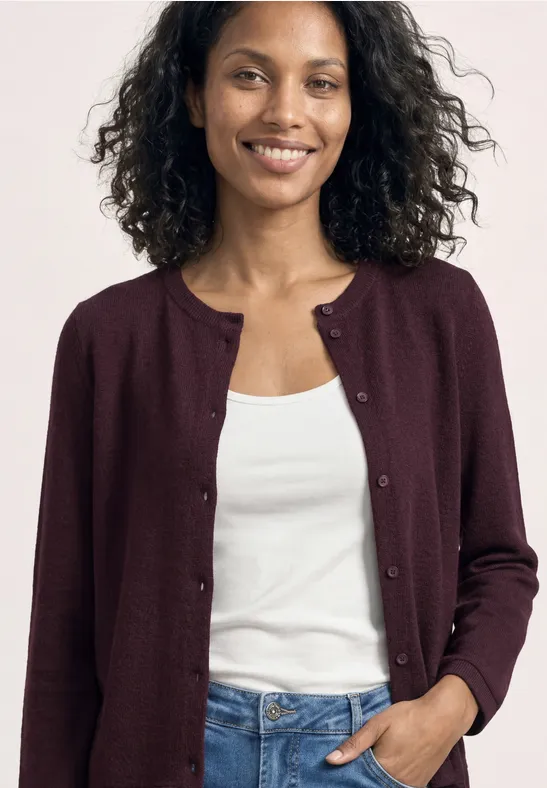 Vrouw in bordeauxrode cardigan, witte top en spijkerbroek, krullend haar, hand in zak.