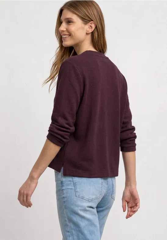 Vrouw met krullend haar draagt een bordeauxvest en blauwe jeans, zijaanzicht.