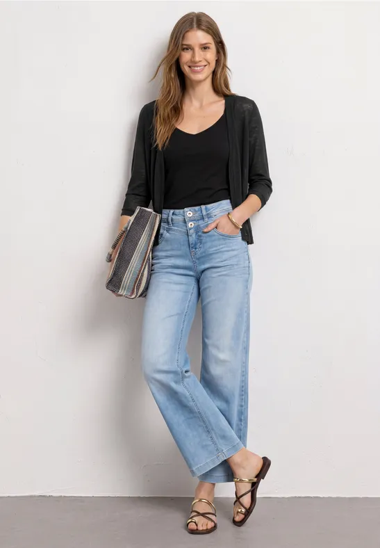 Vrouw in zwarte cardigan, zwarte top, lichtblauwe jeans en sandalen, met gestreepte clutch.