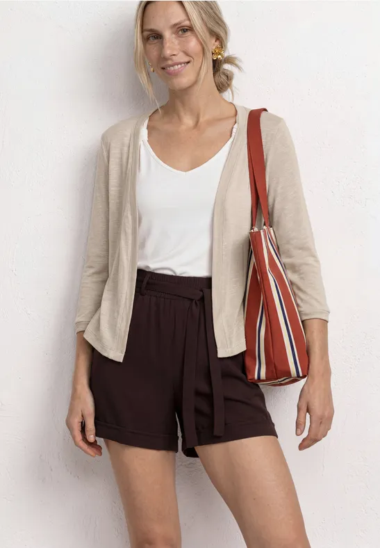 Frau trägt weißen Top, beigen Cardigan, braune Shorts und eine gestreifte Tasche.