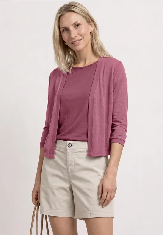 Blonde Frau in rosa Shirt und Cardigan mit beigen Shorts und passender Tasche vor weißem Hintergrund.