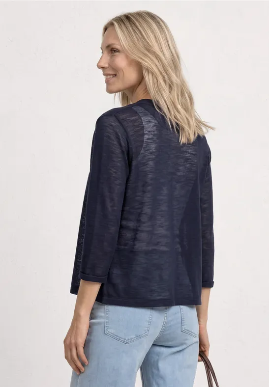 Vrouw van achteren in een marineblauw licht vest met driekwarts mouwen en een lichte spijkerbroek, met tas in de hand.