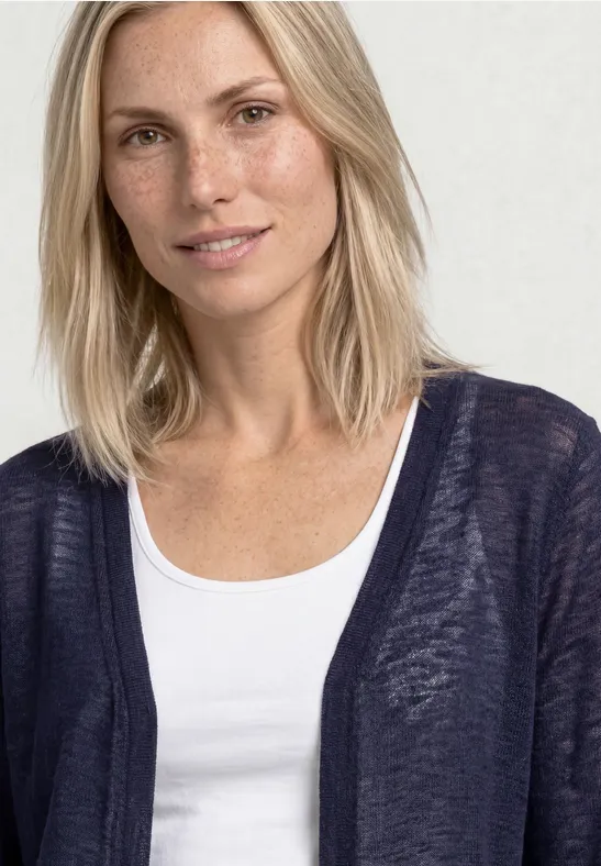Een vrouw met halflang blond haar draagt een wit hemdje en een donkerblauwe cardigan.