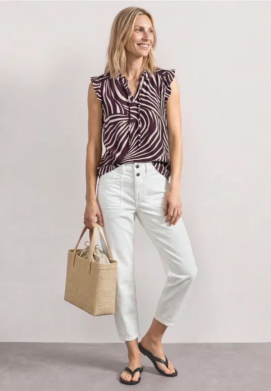 Vrouw in zebraprint top, witte jeans, zwarte slippers en rieten tas voor neutrale achtergrond.