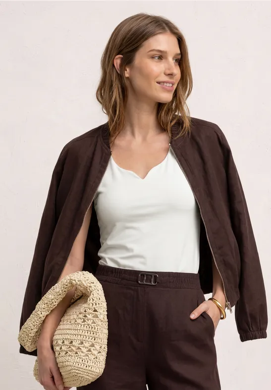 Frau in weißem Top, braunem Blazer und passender Hose hält eine geflochtene Strohtasche.