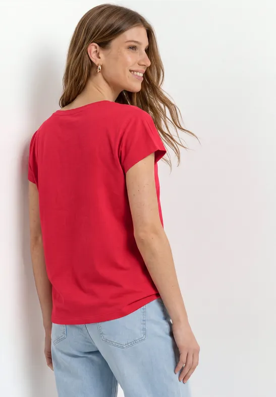 Vrouw van achteren in een rood t-shirt en lichte spijkerbroek tegen witte achtergrond.