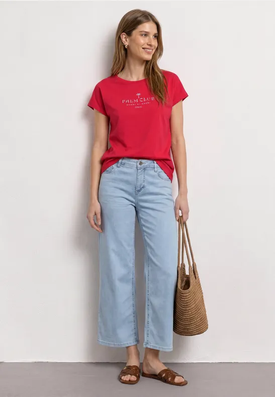 Een vrouw in een rood T-shirt, lichtblauwe jeans, sandalen en rieten tas voor een witte muur.