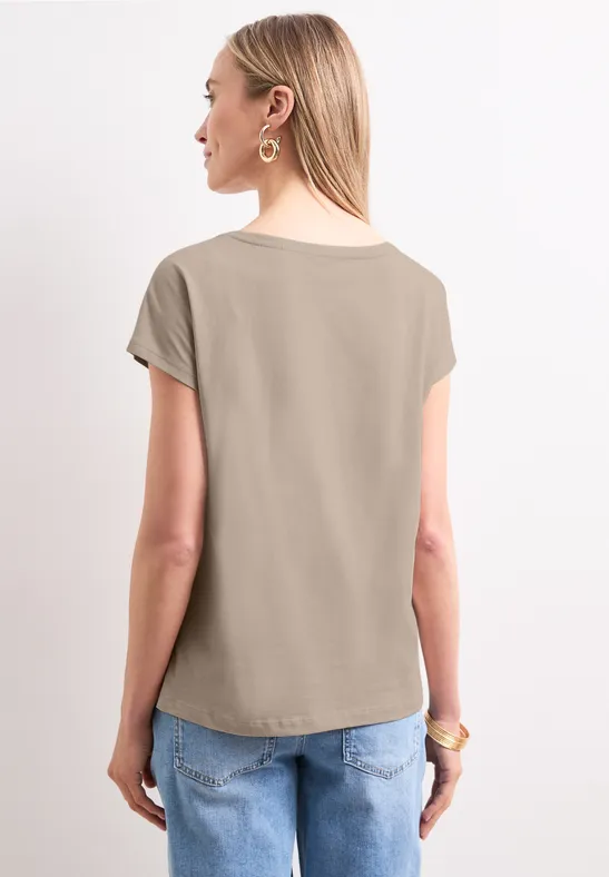 Vrouw staat met de rug naar de camera, draagt een kaki t-shirt en een blauwe spijkerbroek.