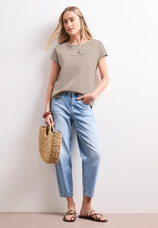 Vrouw in beige T-shirt, lichtblauwe jeans, sandalen en rieten tas.