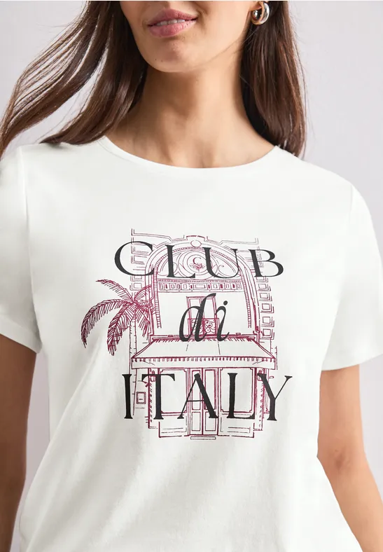 Frau trägt weißes T-Shirt mit 'Club di Italy'-Aufdruck, pastellfarbene Buchstaben und Palmenmotiv.