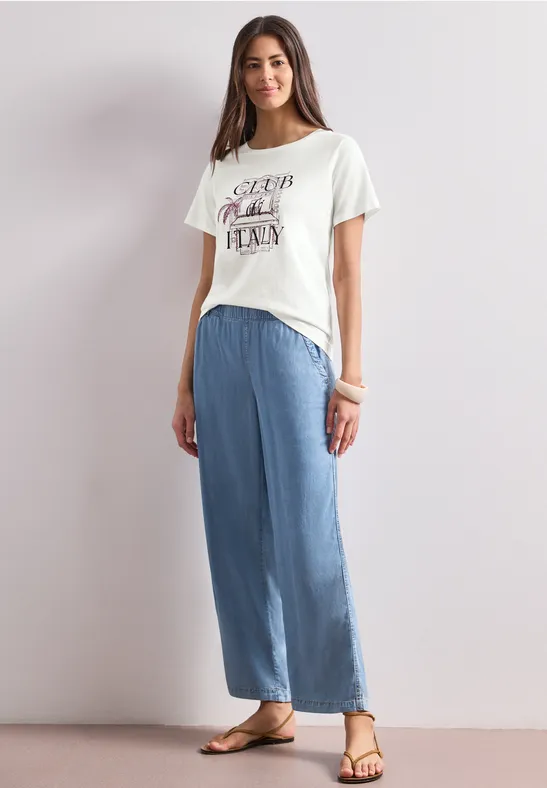 Frau im weißen T-Shirt und weiten blauen Hosen, stehend vor heller Wand in Sandalen.
