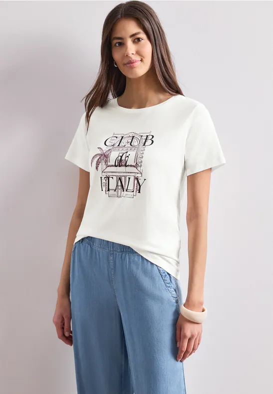 Eine Frau trägt ein weißes T-Shirt mit Aufdruck und lockere, hellblaue Hose vor neutralem Hintergrund.