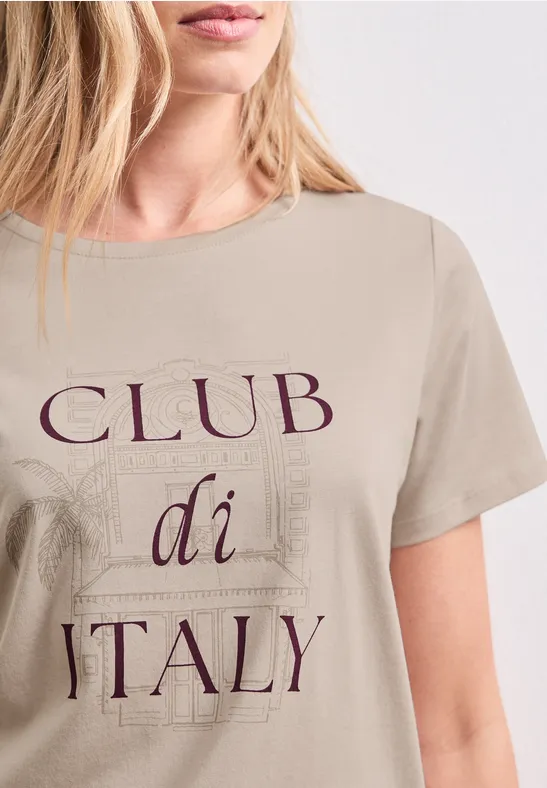 Een vrouw draagt een beige T-shirt met de tekst "CLUB di ITALY" en subtiele architecturale motieven.
