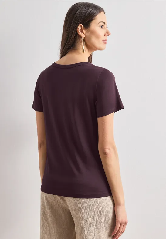 Frau von hinten in dunkelviolettem T-Shirt und hellen, beigen Hosen vor weißem Hintergrund.