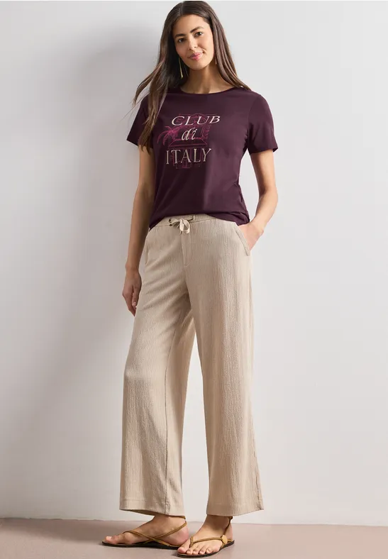 Eine Frau steht in minimalistischem Umfeld, trägt ein lila T-Shirt mit 'Club de Italy', beige weite Hose und Sandalen.