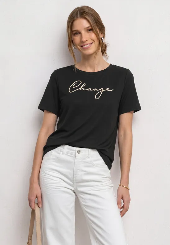 Vrouw met zwart T-shirt 'Change' en witte broek, draagt een tas, voor een lichte achtergrond.
