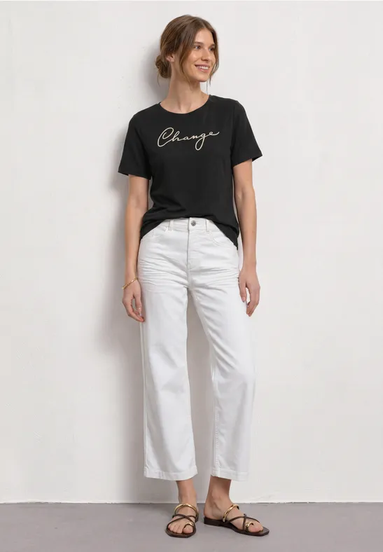 Een vrouw in een zwart T-shirt, witte broek en platte sandalen voor een lichte achtergrond.