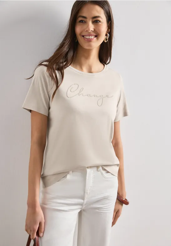 Frau trägt beiges T-Shirt mit 'Challenge'-Schriftzug und weiße Hose, lässiger Casual-Look.