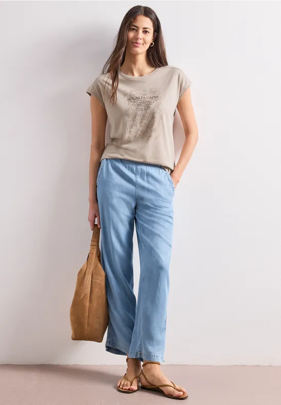 Frau in lässigem Outfit: beiges T-Shirt, weite blaue Jeans, Sandalen und Handtasche.