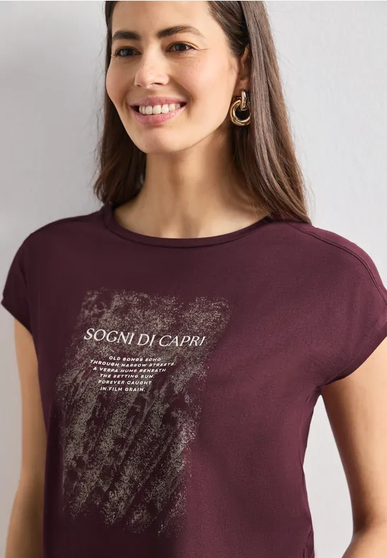 Frau trägt burgunderrotes T-Shirt mit weißem Grafikdruck und Schriftzug „SOGNI DI CAPRI“, lächelnd.