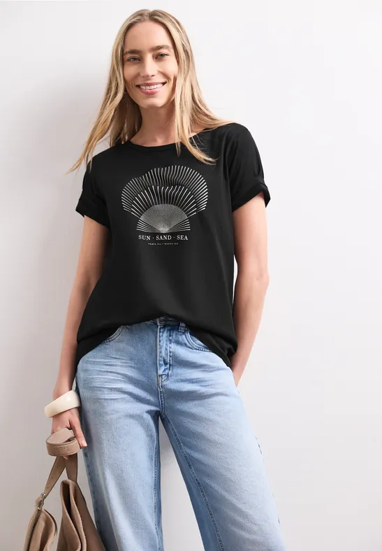 Vrouw draagt zwart bedrukt T-shirt, lichtblauwe jeans en houdt een bruine tas vast.