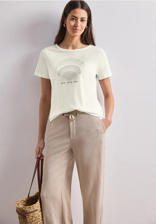 Frau in weißem T-Shirt mit Muster, beiger Hose und geflochtener Korbhandtasche.