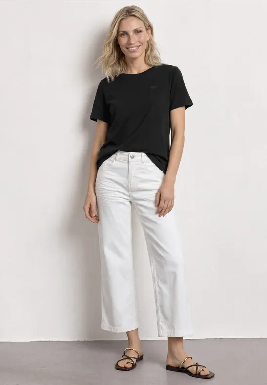 Frau mit schwarzem T-Shirt, weißen, weiten, knöchellangen Jeans und schwarzen Sandalen vor weißem Hintergrund.