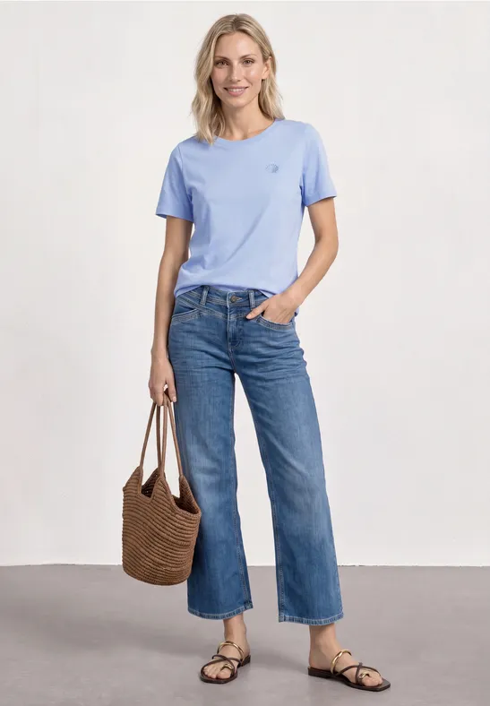 Frau in hellblauem T-Shirt, weiter Jeans, Sandalen und Korbtasche.