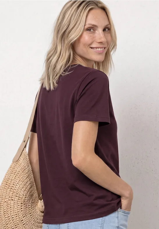 Frau mit blondem Haar, trägt dunkelviolettes T-Shirt und Korbtasche, seitliche Rückansicht.