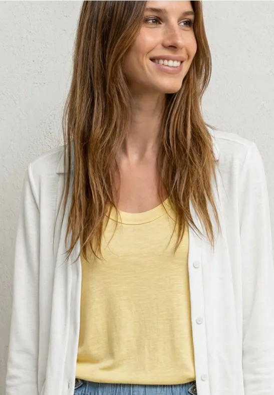 Vrouw met lang haar draagt een geel hemdje en een witte cardigan voor een lichte achtergrond.