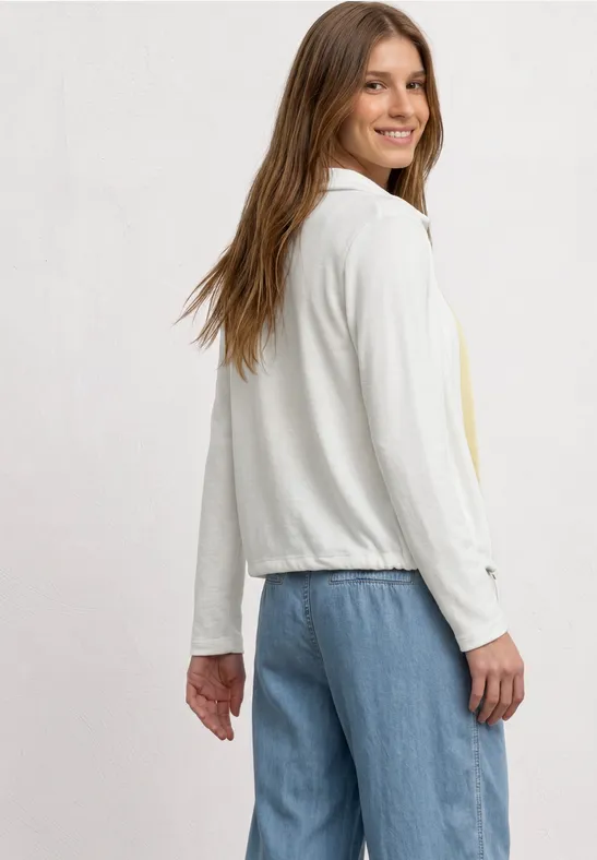 Vrouw in zijaanzicht met een lichte witte jas met lange mouwen en een blauwe spijkerbroek.