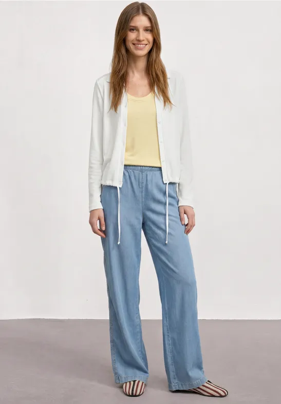 Een vrouw draagt een witte jas, geel shirt, losse lichtblauwe broek en gestreepte sandalen tegen effen achtergrond.