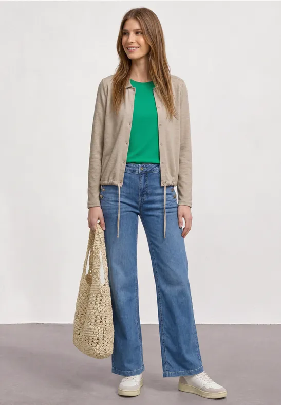 Junge Frau trägt grünes T-Shirt, helle Jacke, blaue Jeans, weiße Sneaker und eine gehäkelte Tasche.