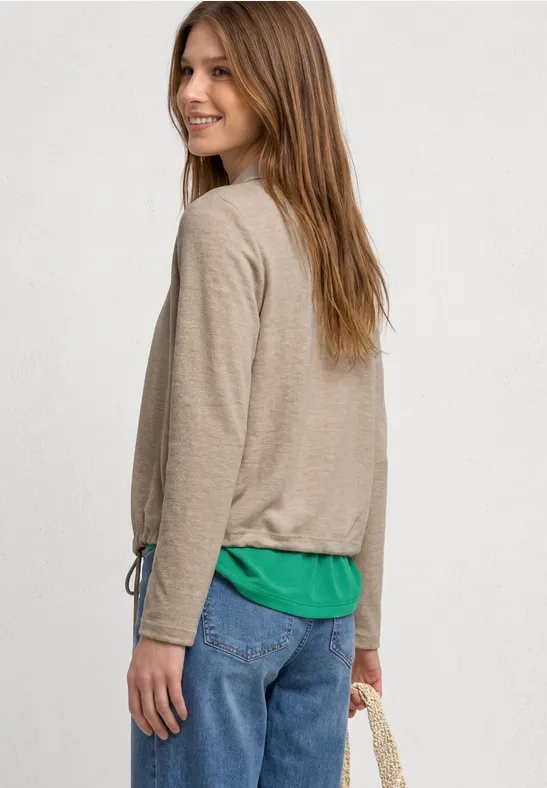 Frau von hinten in beigem Longsleeve, grünem Top, Jeans und mit umgehängter Tasche.