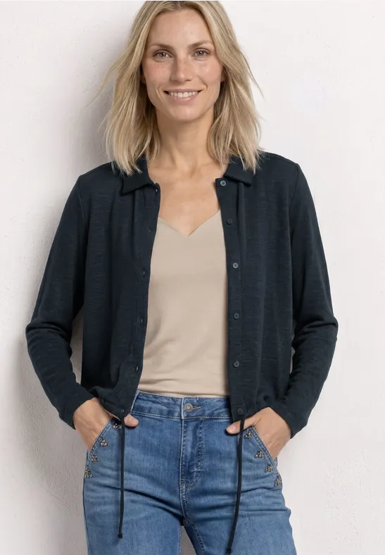 Eine Frau mit hellblondem Haar trägt eine Jeans, ein beiges Top und eine dunkle Strickjacke vor weißem Hintergrund.