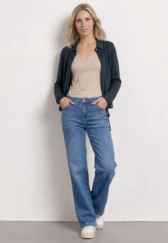 Frau vor weißer Wand mit beigem Top, grauer Strickjacke, gerader Jeans und weißen Sneakern.