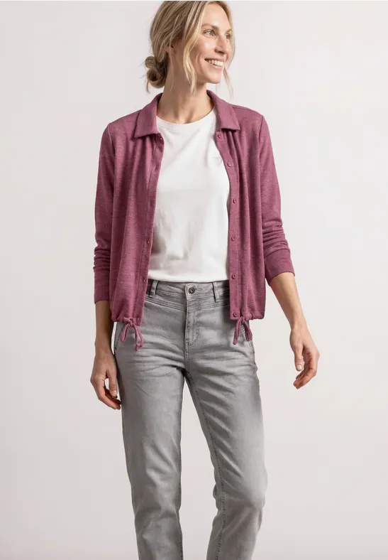 Frau in grauen Jeans, weißem T-Shirt und offener rosa Strickjacke vor hellem Hintergrund
