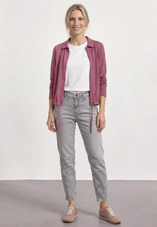 Junge Person trägt weißes T-Shirt, karierte rosa Hemdjacke, graue Jeans und rosa Sneaker vor neutralem Hintergrund.