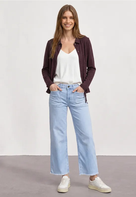 Eine Frau steht vor neutralem Hintergrund und trägt weiße Bluse, weite hellblaue Jeans, Cardigan und Sneakers.