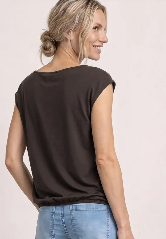 Frau von hinten im dunkelbraunen T-Shirt und hellen Jeans vor hellem Hintergrund.
