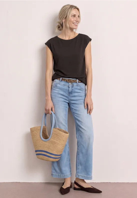 Frau lehnt an heller Wand, trägt schwarzes T-Shirt, weite Jeans, Flechtfrisur und eine Stroh-Tasche.