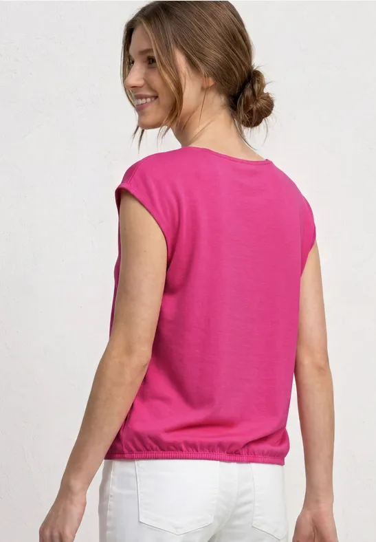 Een vrouw met opgestoken haar draagt een roze top en witte broek, ruggezicht tegen een lichte achtergrond.