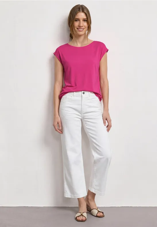 Een vrouw draagt een roze top, witte broek en sandalen, staand voor een lichte achtergrond.