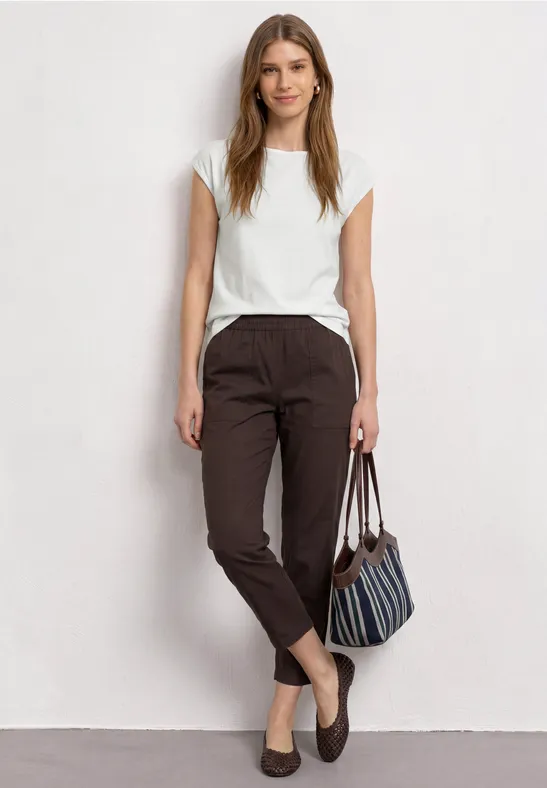 Een vrouw in casual kleding staat voor een witte muur, met witte top, bruine broek en tas.