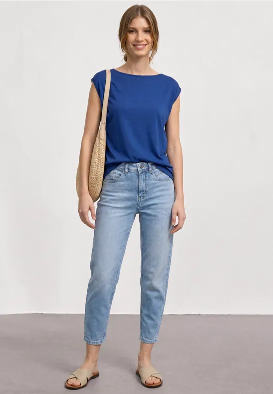 Vrouw met blauw mouwloos topje, lichtblauwe jeans, sandalen en strooien tas voor neutrale achtergrond.