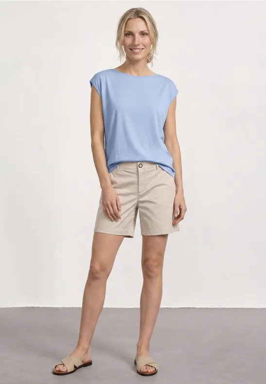 Vrouw in lichtblauw shirt en beige korte broek, staand voor een neutrale achtergrond.