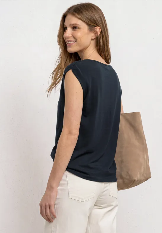 Seitliche Aufnahme einer Frau mit schwarzem ärmellosem Top, weißen Hosen und brauner Tasche.