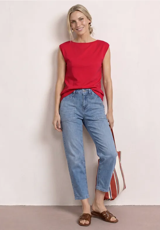 Frau in rotem Top, hellblauen Jeans, braunen Sandalen und gestreifter Strandtasche vor neutralem Hintergrund