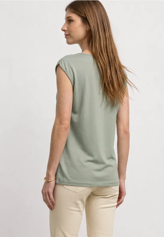 Vrouw van achteren in een loszittend groen shirt zonder mouwen en een beige broek tegen een lichte achtergrond.